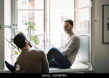 Happy gay man talking to boyfriend while sitting on windowsill at home Stock Photo