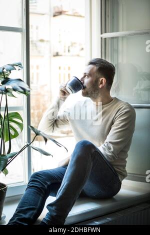 Thoughtful young man drinking coffee while sitting on window sill Stock Photo