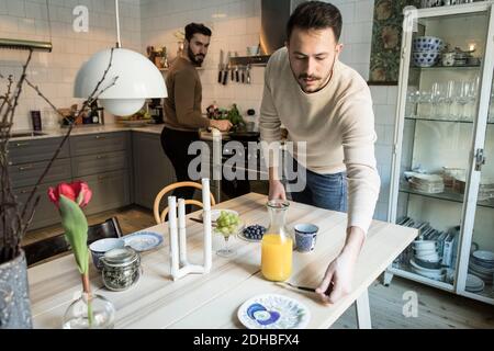 Young man setting table while partner working in background at kitchen Stock Photo