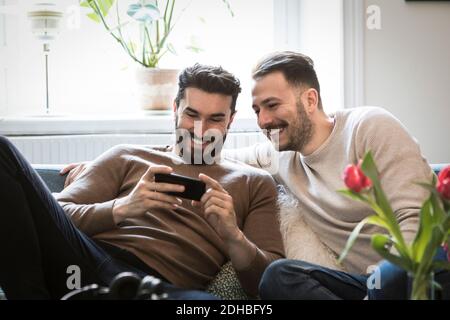 Smiling young men looking at smart phone while sitting on sofa at home Stock Photo