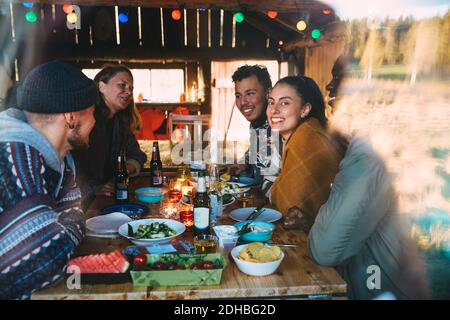 Male and female friends talking while enjoying meal on table in cottage seen through window Stock Photo