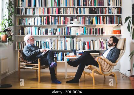 Therapist communicating with man while sitting by book shelf at home office Stock Photo