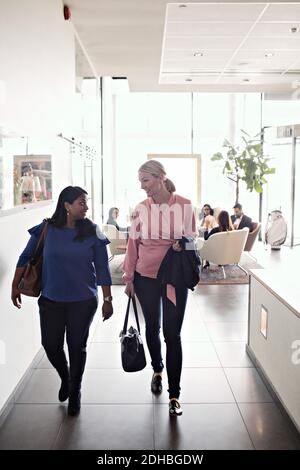 Businesswomen talking while leaving office with colleagues sitting in background Stock Photo