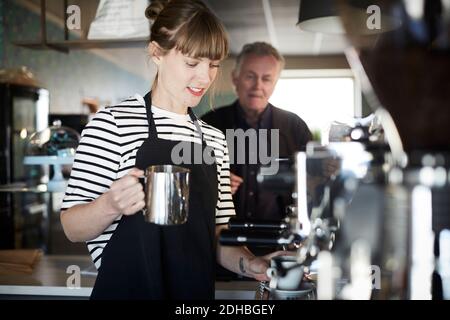 Female barista holding pitcher while making coffee with customer standing in background at cafe Stock Photo