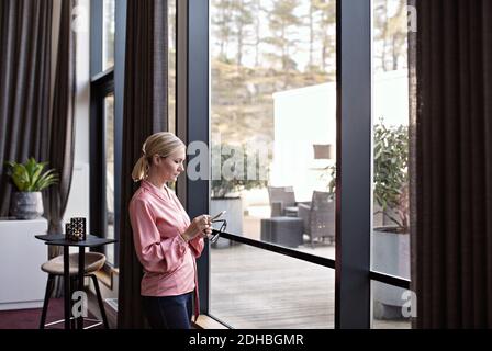 Businesswoman using phone while standing by window in office Stock Photo