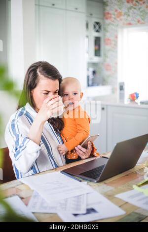 Working businesswoman drinking coffee while carrying yawning daughter in dining room Stock Photo