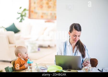 Baby girl looking at busy working mother while sitting at table in home office Stock Photo