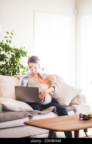 Multi-tasking mother using laptop while caring baby girl on sofa at home office Stock Photo