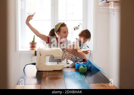 Female tailor taking selfie with daughter through smart phone while sitting on table at home Stock Photo