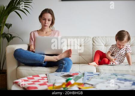 Fashion designer looking at laptop while daughter using digital tablet on sofa at home Stock Photo