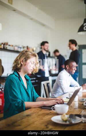 Serious female IT hacker using laptop while colleagues talking in background at creative workplace Stock Photo