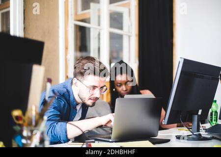 Young computer expert coding in laptop while working with female colleague at workplace Stock Photo