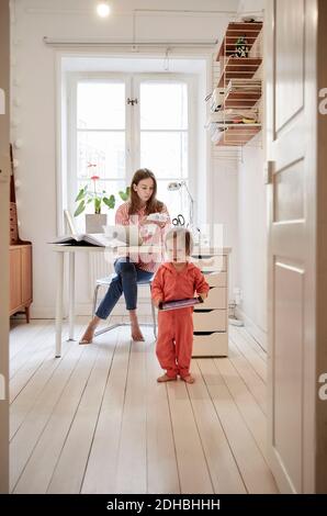 Girl holding digital tablet while woman working on financial bills at table seen through doorway Stock Photo
