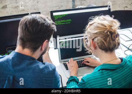Rear view of female IT expert coding in laptop while sitting by male hacker at workplace Stock Photo