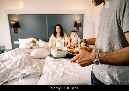 Father holding breakfast with family sitting on bed in background Stock Photo