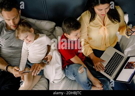 Directly above shot of parents using technologies while sitting with children on bed Stock Photo