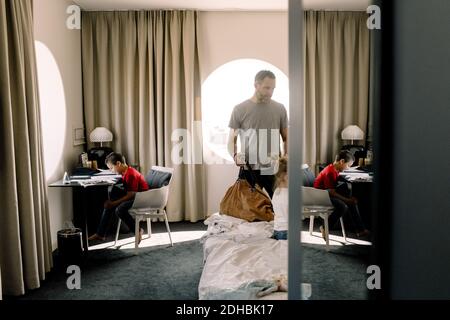 Man holding bag by bed while boy sitting at table in hotel Stock Photo