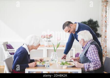Son showing mobile phone to mother and father having lunch at table in nursing home Stock Photo
