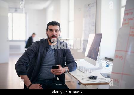 Portrait of male engineer holding mobile phone while sitting at computer desk in office Stock Photo