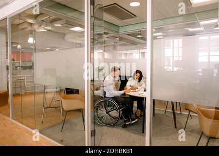 Businessman sitting on wheelchair while working with female colleague in board room at work place seen through doorway Stock Photo