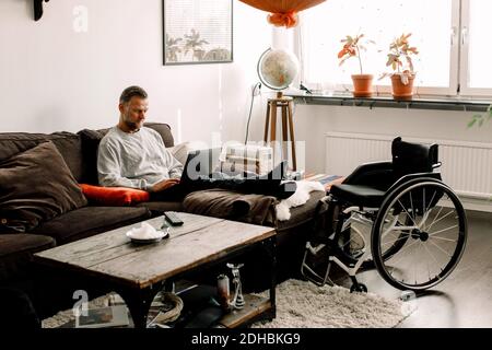 Full length of disabled mature man using laptop while sitting on sofa in living room at home Stock Photo