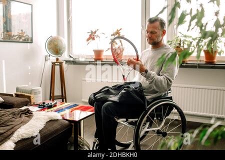 Thoughtful mature man holding tennis racket while sitting on wheelchair at home Stock Photo
