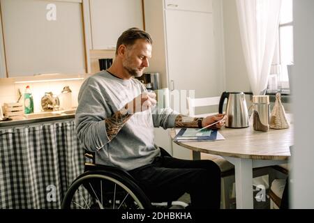 Tattooed mature man reading mail while holding coffee cup on wheelchair at home Stock Photo