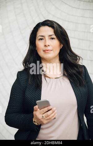 Portrait of confident businesswoman holding smart phone standing against wall at office Stock Photo