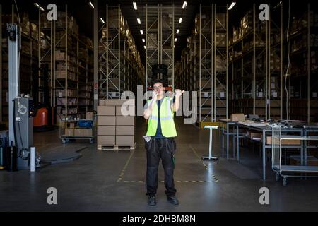Full length of young male warehouse worker gesturing while using walkie-talkie at storage room Stock Photo