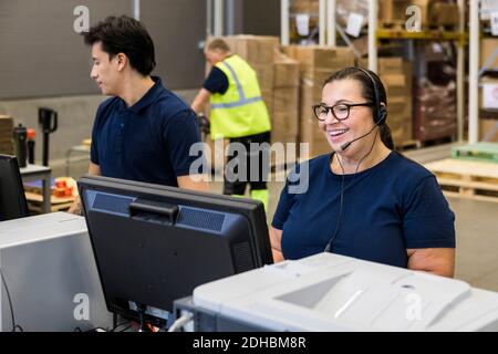 Smiling mature female customer service representative talking through headset while standing by coworker in distribution Stock Photo