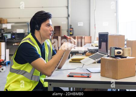 Side view of confident young male customer service representative talking through headset while sitting at desk in wareh Stock Photo