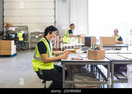 Young customer service representative talking through headset while coworkers at warehouse Stock Photo