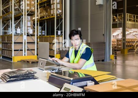 Confident young male customer service representative using laptop while sitting at desk in warehouse Stock Photo