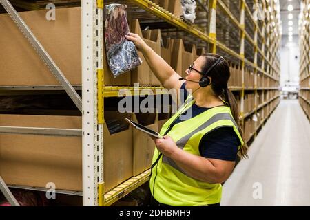 Mature female worker with digital tablet checking merchandise on rack while talking through headset at distribution ware Stock Photo