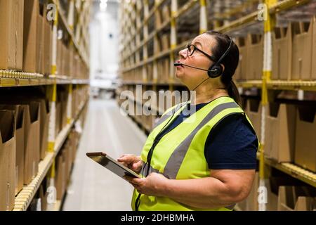 Mature female worker with digital tablet looking up while talking on headset at distribution warehouse Stock Photo