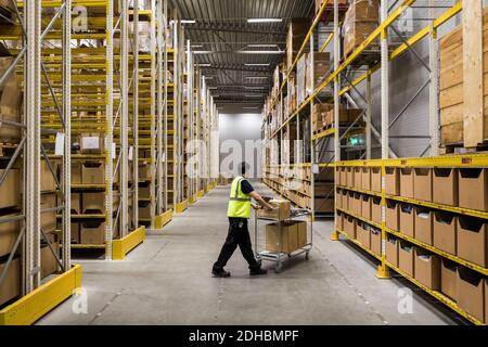 Full length of young warehouse worker pushing cart on aisle in industrial building Stock Photo