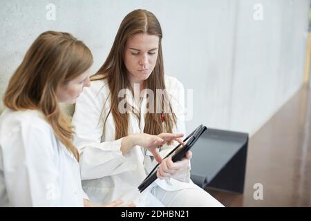 Confident female doctors discussing over digital tablet while sitting in corridor at hospital Stock Photo