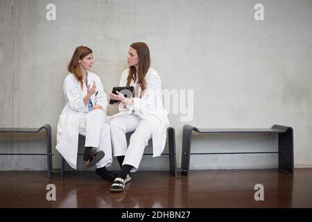 Full length of confident female doctors discussing over digital tablet while sitting against wall at hospital Stock Photo