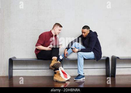 Full length of young male students using technologies while sitting on bench against wall in university Stock Photo
