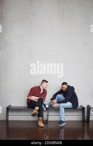 Full length of happy male students with technologies sitting on bench against wall in university Stock Photo