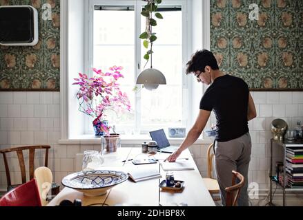 Rear view of young man reading book on table while standing at home Stock Photo