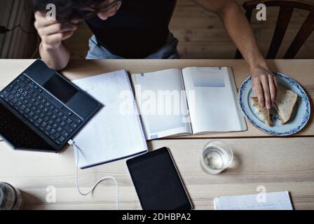 High angle view of dedicated young man doing homework while having sandwich at table Stock Photo