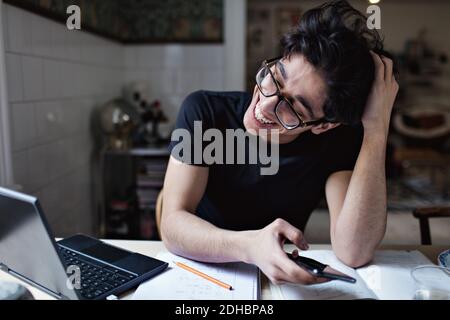 Cheerful social media addicted young man using mobile phone while doing homework Stock Photo