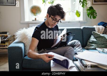 Serious young man doing homework while sitting on sofa with mobile phone and laptop at home Stock Photo