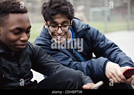 Smiling young man looking at friend's mobile phone against playing field in city during winter Stock Photo
