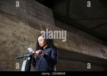 Low angle view of teenage girl with electric push scooter using mobile phone by wall below bridge Stock Photo