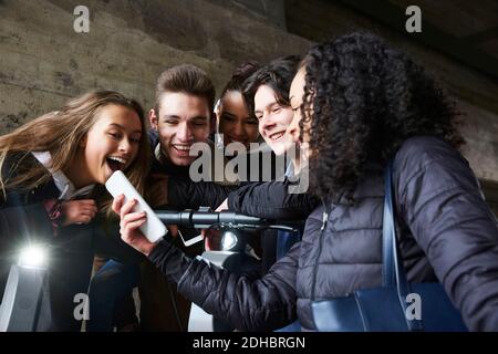 Teenage girl showing mobile phone to happy male and female friends Stock Photo