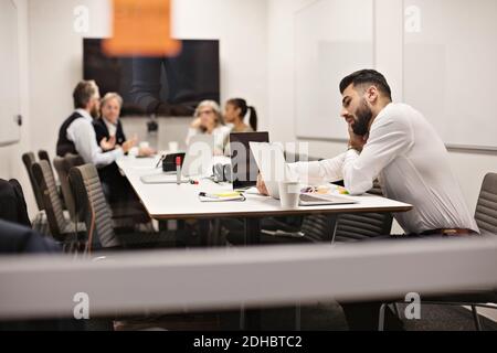 Young businessman talking on phone while using laptop at conference table with colleagues in background Stock Photo