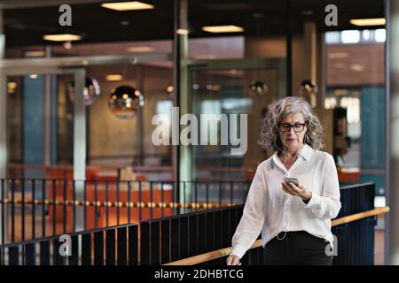 Female senior professional with headphones using smart phone while walking in atrium at office Stock Photo