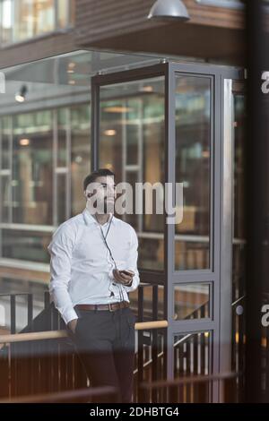 Young thoughtful businessman with headphones using smart phone while standing at office Stock Photo